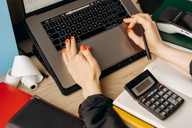 A woman with red nails using a laptop and calculator on a wooden desk. A consistent way to research rival accounting firms, from entity checks to pricing