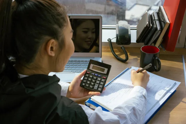Woman using a calculator at a desk with a laptop and notepad, involved in office work. Accounting services market outlook: three 2027 scenarios